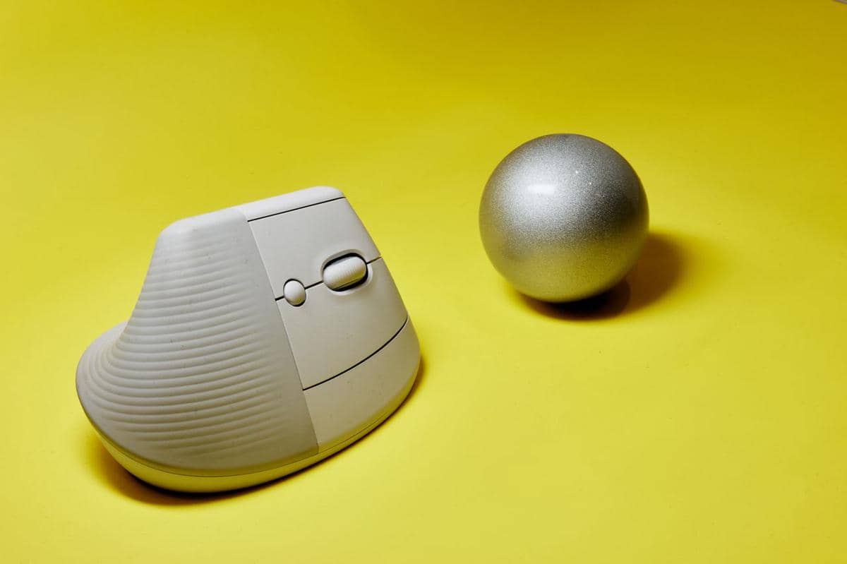 Sebuah vertical mouse berwarna abu-abu muda berdiri di atas permukaan kuning dengan bola perak di sebelah kanannya.