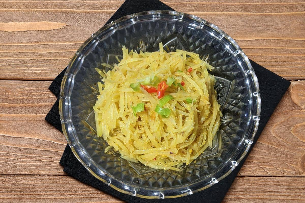 Oseng pepaya muda berbumbu kuning disajikan dalam mangkuk kaca di atas alas kain hitam di meja kayu.