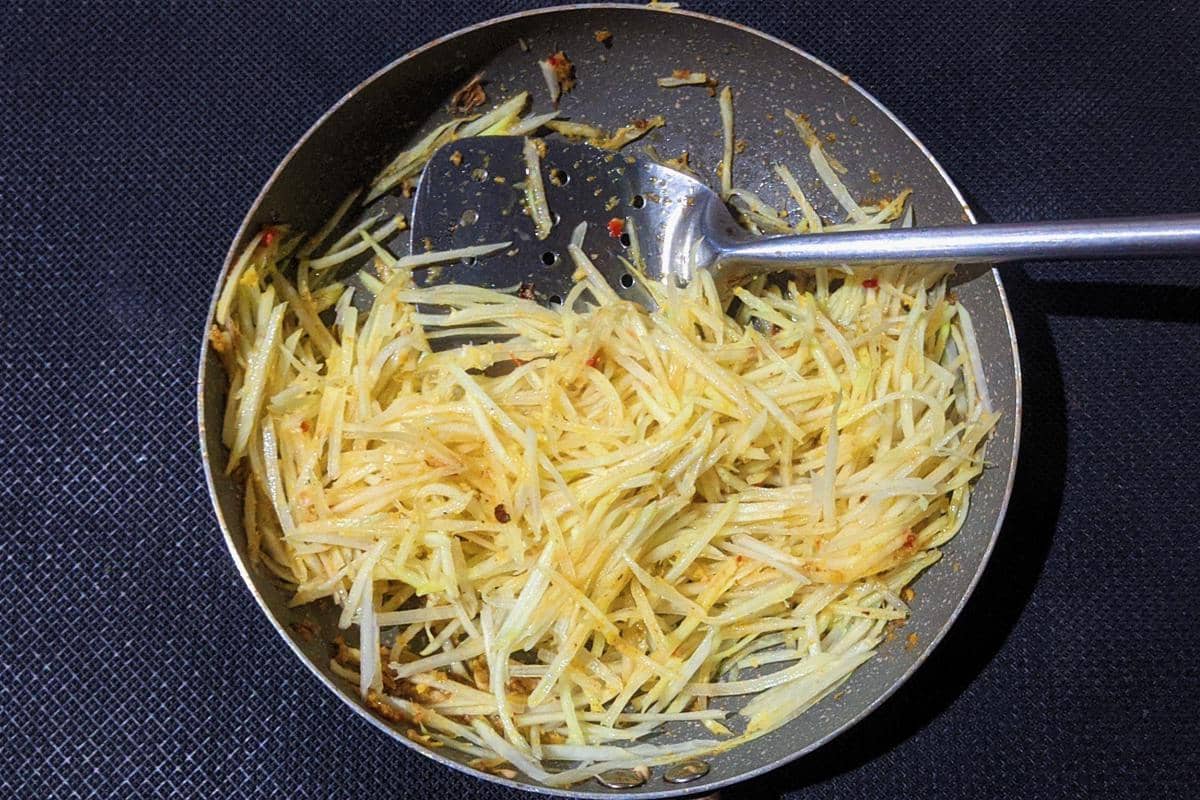 Tumis pepaya muda parut dimasak dengan bumbu kuning di dalam wajan bersama spatula logam di atas alas hitam.
