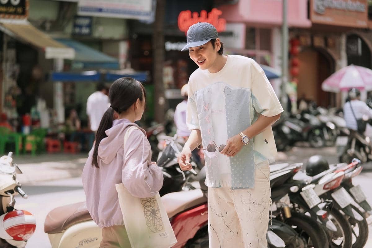 Seorang pria mengenakan topi biru dan kaus terang berbicara dengan seorang wanita berjaket ungu muda di tepi jalan yang ramai dengan deretan motor.