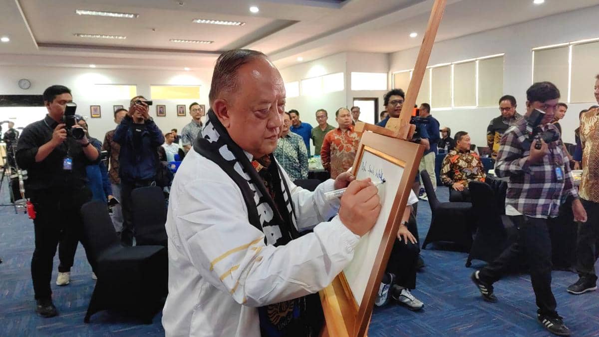 Seorang pria menandatangani kanvas di atas papan kayu saat peluncuran buku Sports Intelligence oleh KONI di UNESA, disaksikan peserta acara.