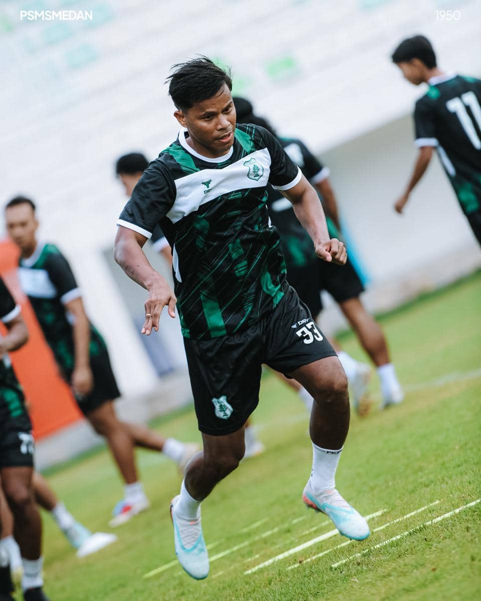 Seorang pemain PSMS Medan berlatih di lapangan rumput dengan mengenakan seragam latihan berwarna hitam dan hijau.