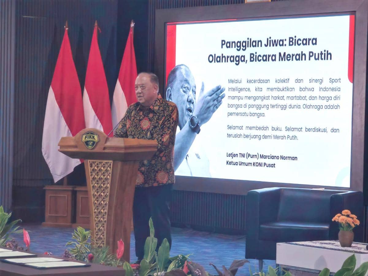 Seorang pembicara berdiri di podium berlogo FIKK dengan latar layar besar bertuliskan pesan tentang olahraga dan tiga bendera Indonesia di belakangnya.