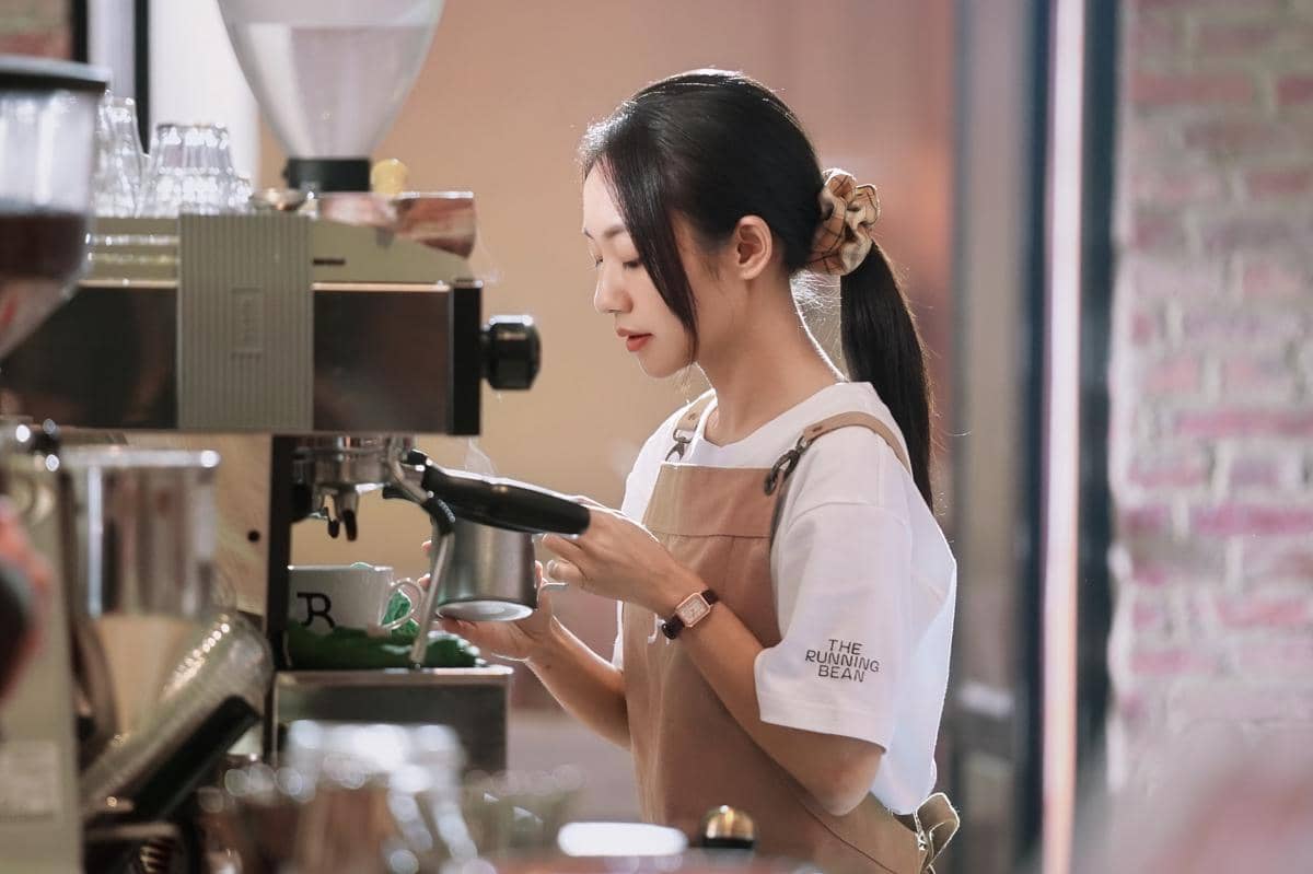 Seorang barista perempuan mengenakan celemek cokelat sedang membuat kopi menggunakan mesin espresso di kedai kopi modern.