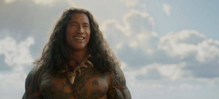 Karakter berambut panjang dan bertato tersenyum di tepi laut dengan latar langit cerah dalam cuplikan film Moana.