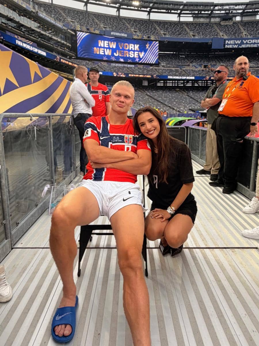 Seorang pria mengenakan jersey tim nasional Norwegia duduk di kursi sementara seorang wanita berjongkok di sampingnya di stadion besar.