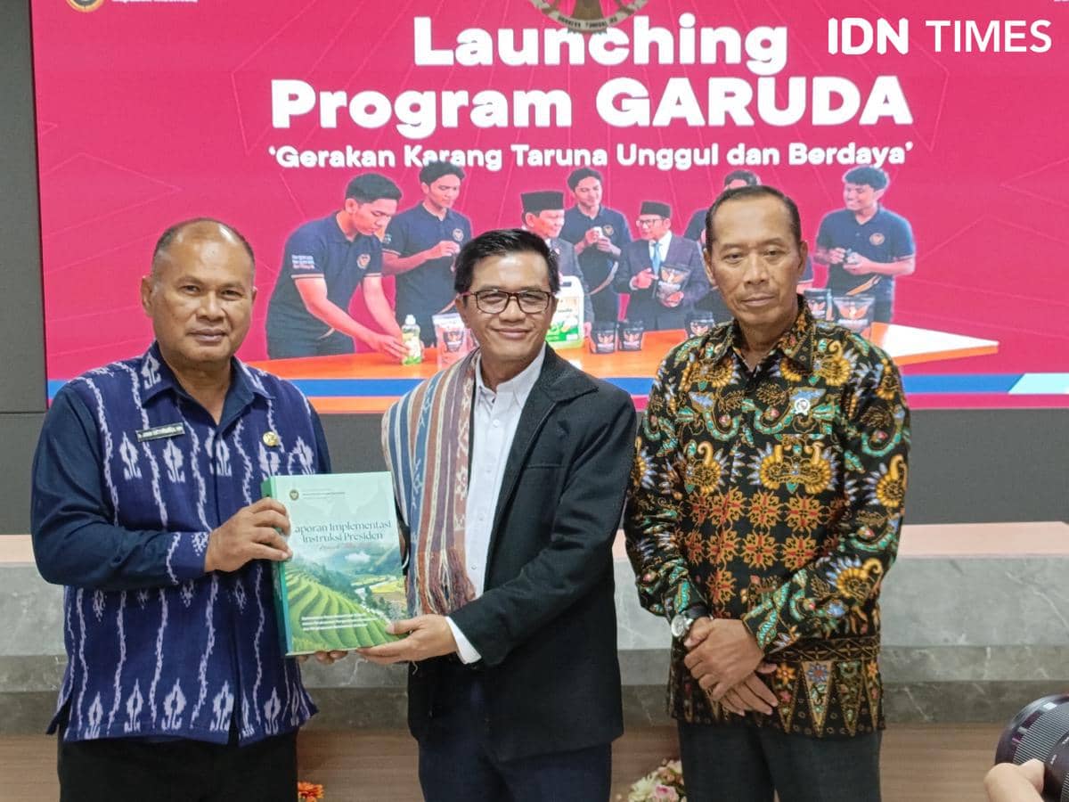 Tiga pria berpose bersama di depan layar bertuliskan Launching Program Garuda di Kota Kupang, salah satunya memegang buku laporan.