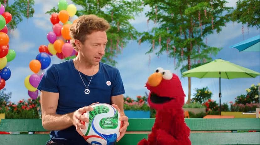 Seorang pria memegang bola sepak sambil berbicara dengan karakter boneka Elmo di taman berwarna cerah dengan balon dan pepohonan.