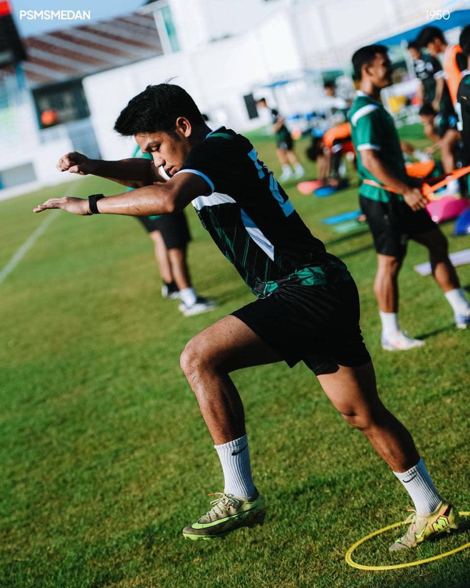 Seorang pemain PSMS Medan berlatih di lapangan rumput dengan mengenakan seragam latihan hijau hitam dan sepatu sepak bola.