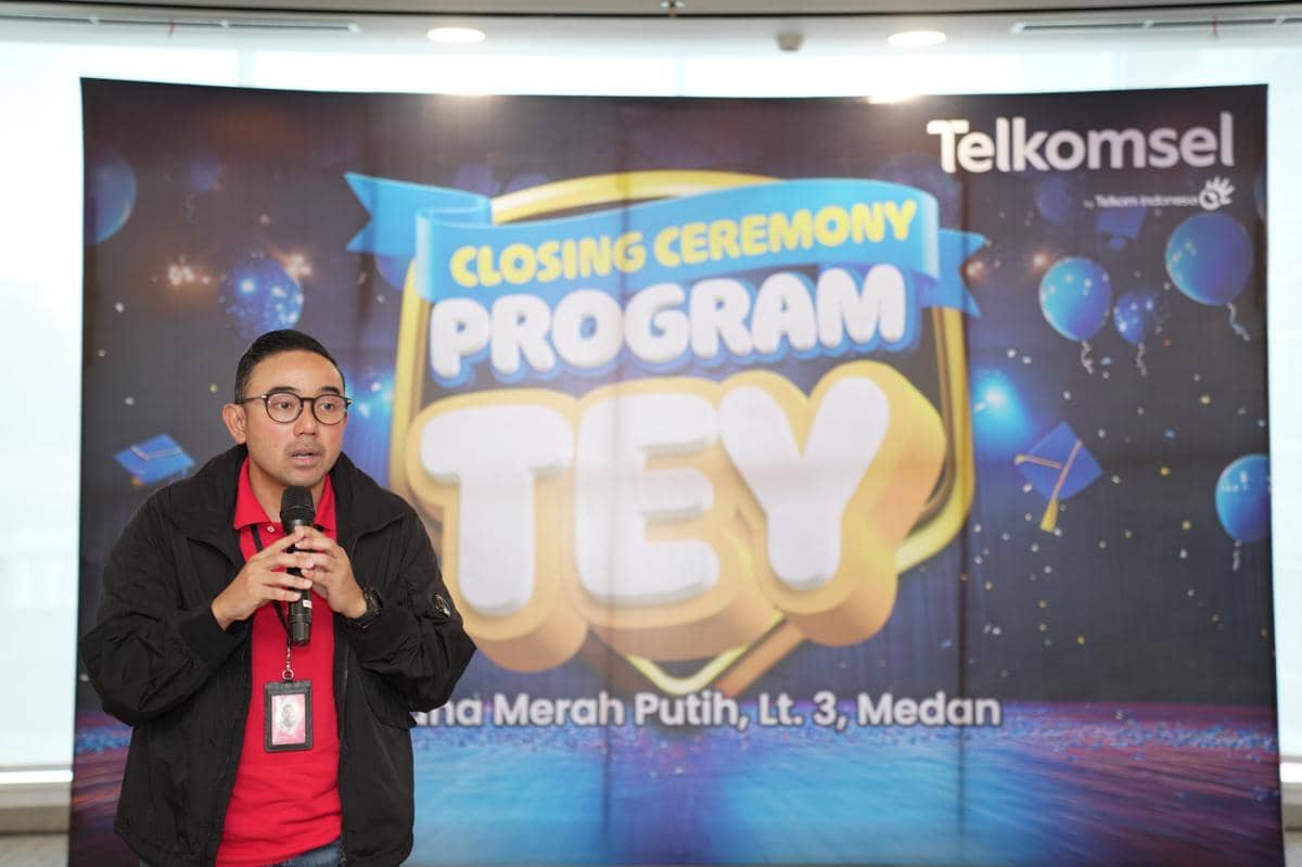 Seorang pria berbicara memegang mikrofon di depan latar belakang bertuliskan Closing Ceremony Program TEY Telkomsel di Graha Merah Putih Medan.