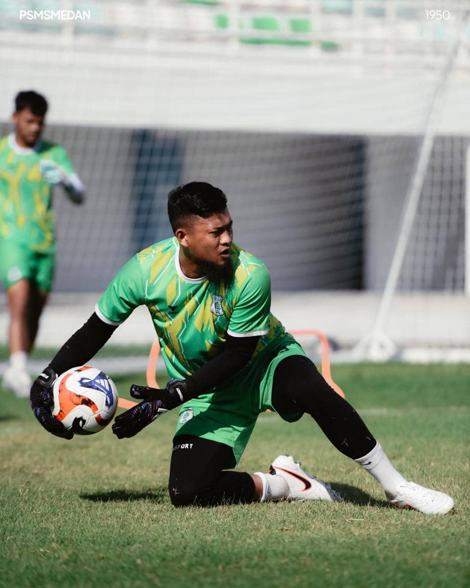 Seorang penjaga gawang PSMS Medan mengenakan seragam hijau sedang berlatih menangkap bola di lapangan sepak bola.