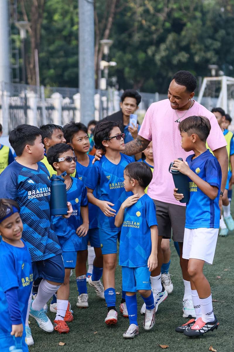 Seorang pelatih berbaju merah muda berbicara dan berinteraksi hangat dengan anak-anak peserta coaching clinic sepak bola di lapangan rumput.