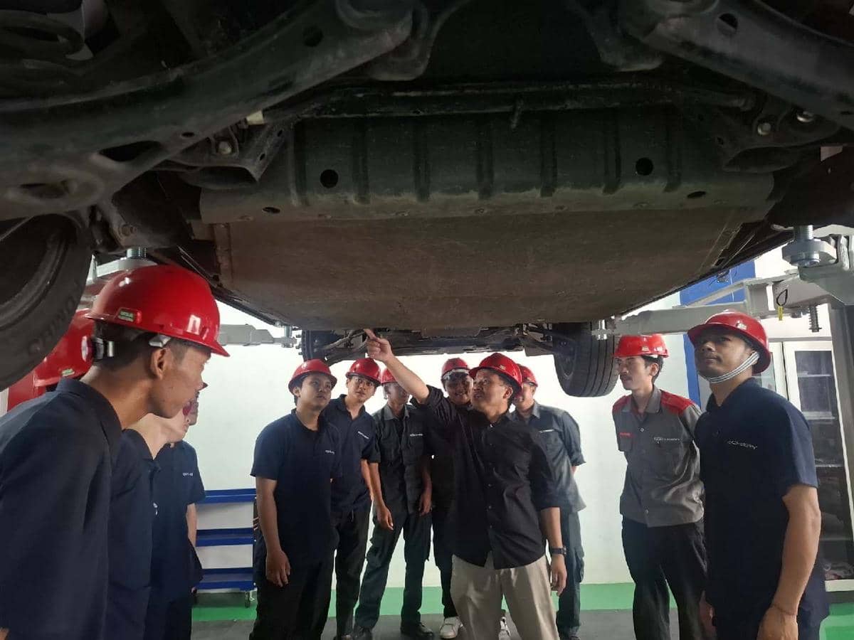 Beberapa teknisi dengan helm merah memeriksa bagian bawah mobil Chery Tiggo Cross yang diangkat di bengkel servis.