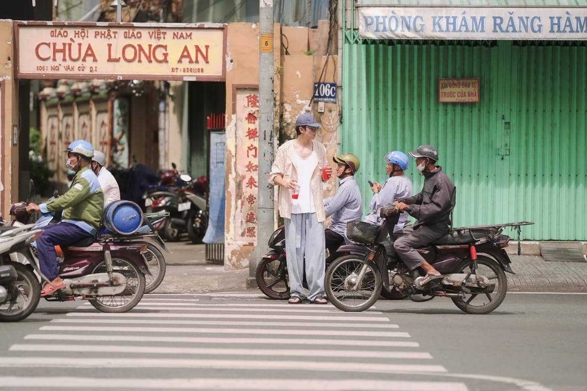 Seorang pria berdiri di tepi jalan sambil memegang minuman, dikelilingi pengendara motor di depan gerbang bertuliskan Chùa Long An di Vietnam.