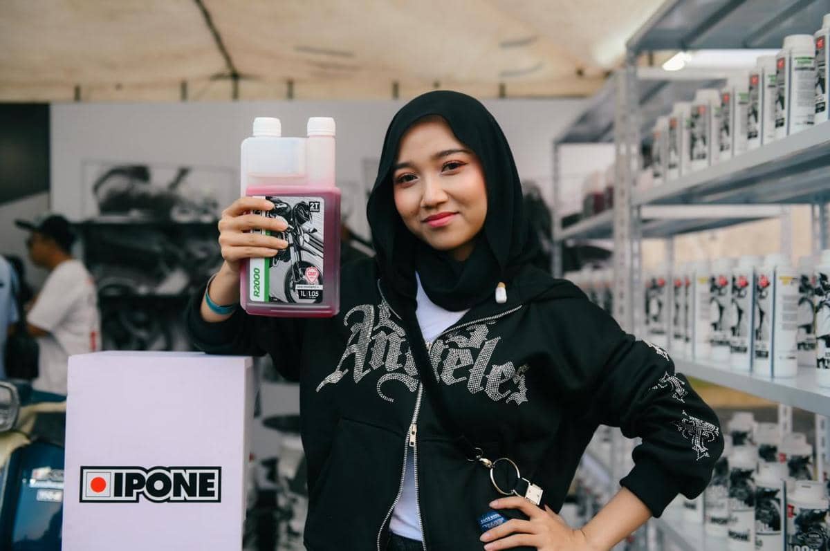 Seorang wanita berhijab memegang botol oli motor merek Ipone di sebuah stan promosi dengan rak berisi produk serupa di latar belakang.