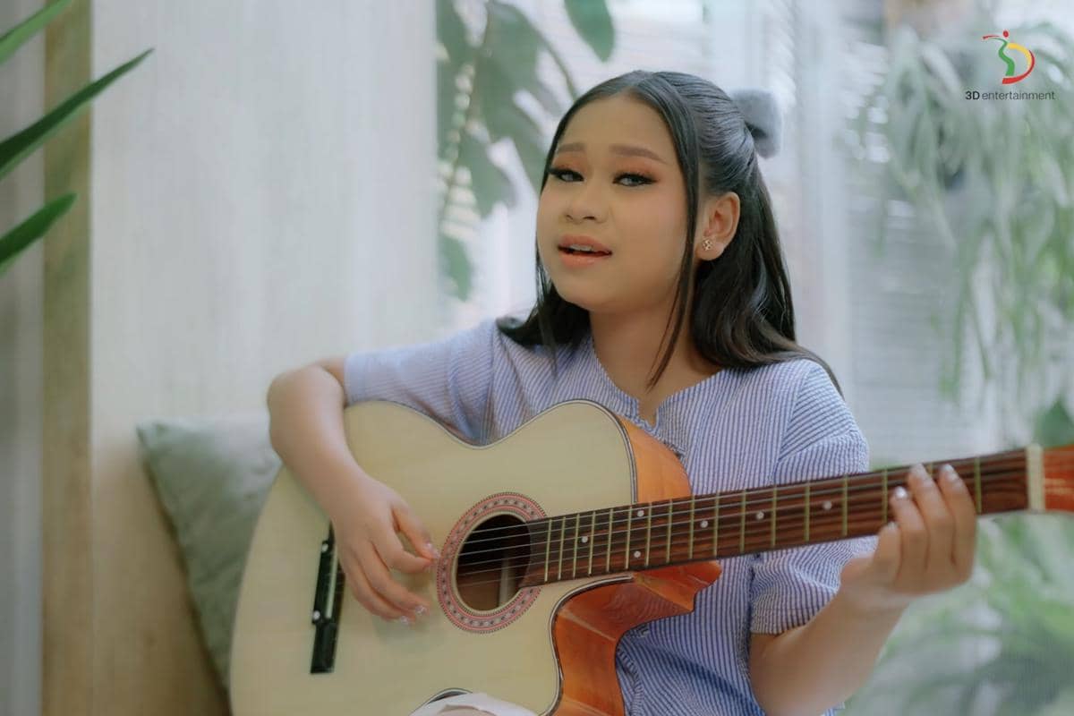 Seorang perempuan muda bernyanyi sambil memainkan gitar akustik di ruangan dengan pencahayaan alami dan latar tanaman hijau.