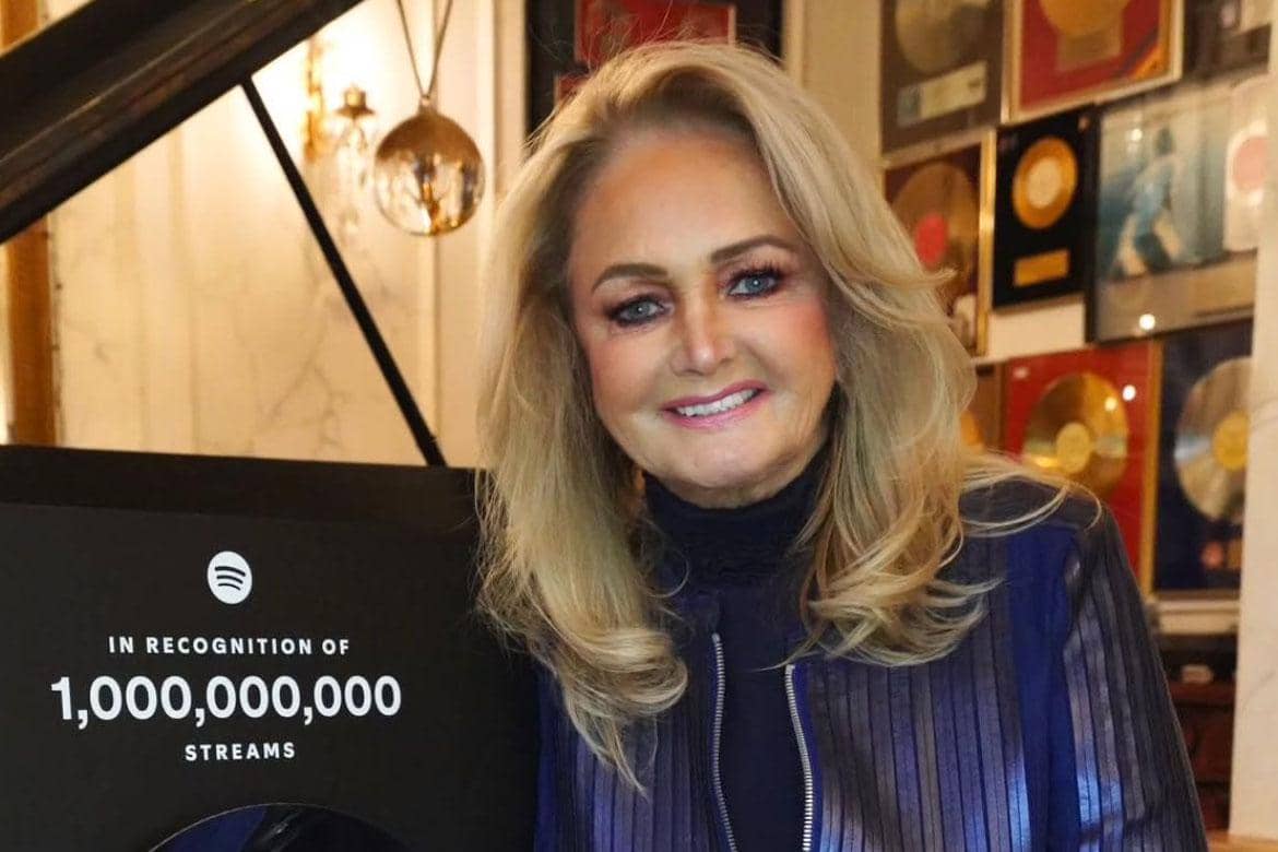 Bonnie Tyler tersenyum di depan penghargaan Spotify atas pencapaian satu miliar streaming di ruangan penuh plakat musik.