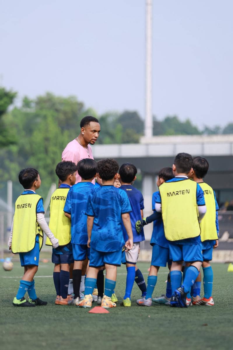 Seorang pelatih memberikan arahan kepada sekelompok anak-anak yang mengenakan seragam sepak bola biru di lapangan hijau terbuka.