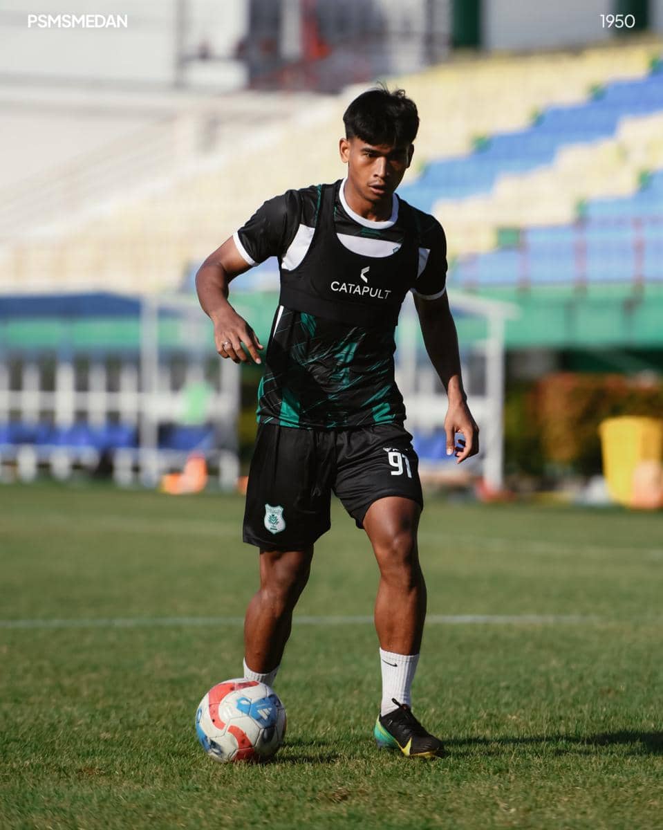Seorang pemain PSMS Medan mengenakan seragam latihan hitam dan hijau sedang menggiring bola di lapangan sepak bola.