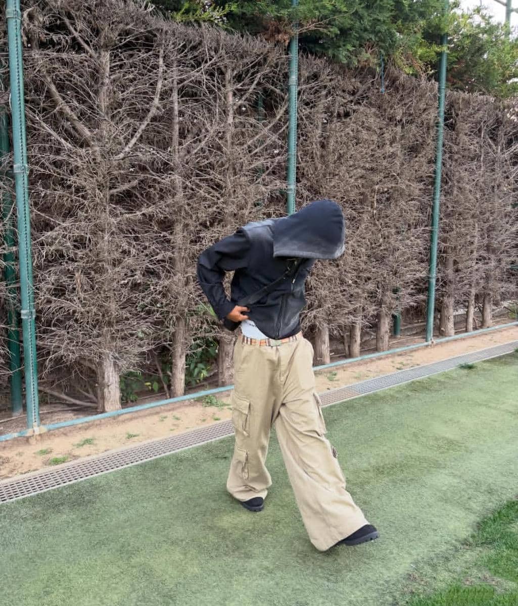 Seseorang mengenakan jaket hitam berkerudung dan celana kargo krem longgar berjalan di area rumput dekat pagar tanaman kering.