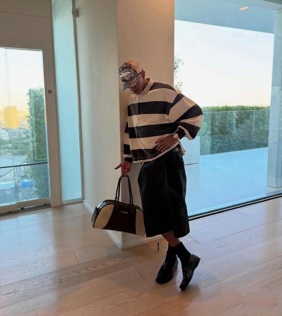 Seorang pria mengenakan sweater garis hitam putih, celana pendek hitam, sepatu hitam, dan topi, sambil memegang tas di ruangan modern.