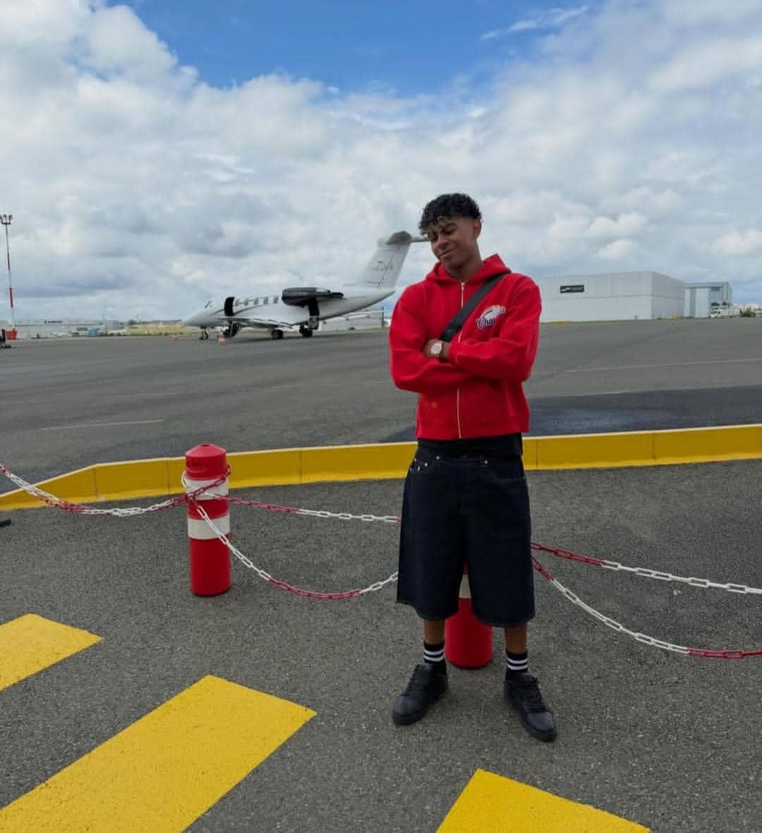 Seorang pria mengenakan jaket merah dan celana pendek denim berdiri di area bandara dengan latar belakang jet pribadi dan langit berawan.