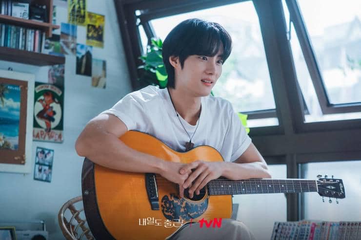 Seorang pria muda duduk di dalam kamar dengan jendela besar sambil memegang gitar akustik berwarna cokelat dan tersenyum santai.