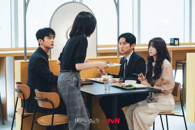 Empat orang sedang duduk di meja makan kantor, dua pria dan dua wanita tampak berbincang serius sambil menikmati hidangan siang.
