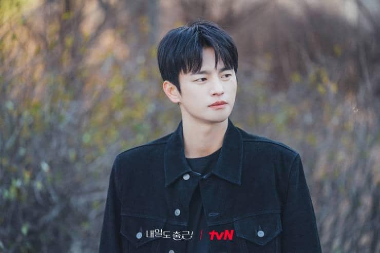 Seorang pria berambut hitam mengenakan jaket hitam berdiri di luar ruangan dengan latar pepohonan dalam adegan drama See You at Work Tomorrow.