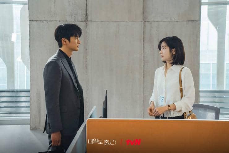 Dua karyawan berdiri saling berhadapan di area kantor modern dalam adegan drama See You at Work Tomorrow dari tvN.