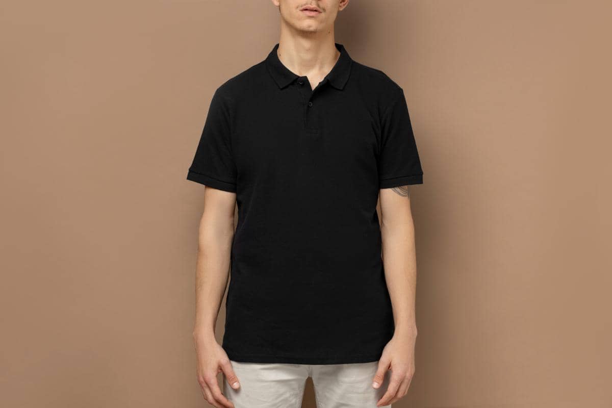Seorang pria mengenakan polo shirt hitam dipadukan dengan celana chino berwarna terang di latar belakang cokelat polos.