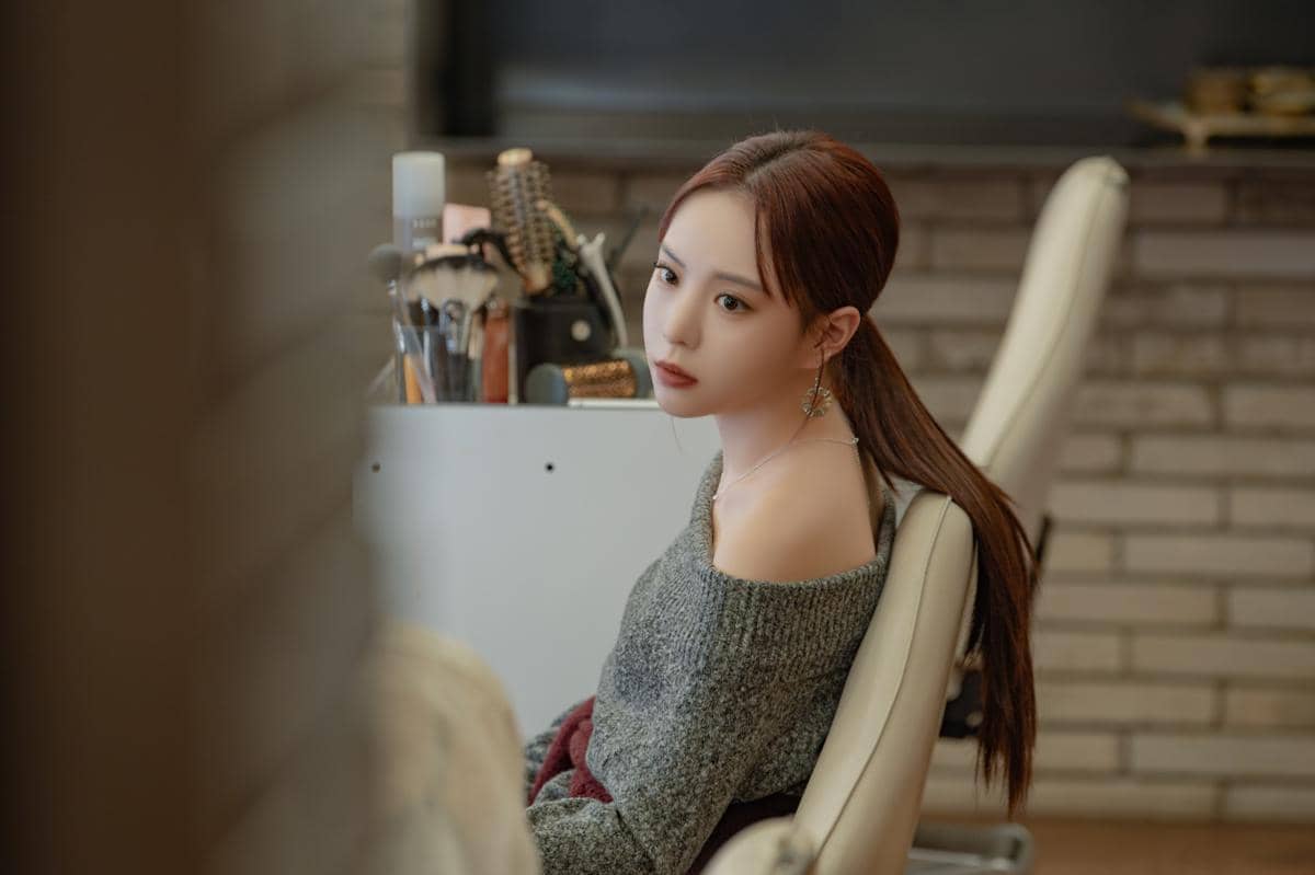 Seorang wanita berambut panjang duduk di kursi salon dengan ekspresi serius, mengenakan sweater abu-abu dan anting panjang.