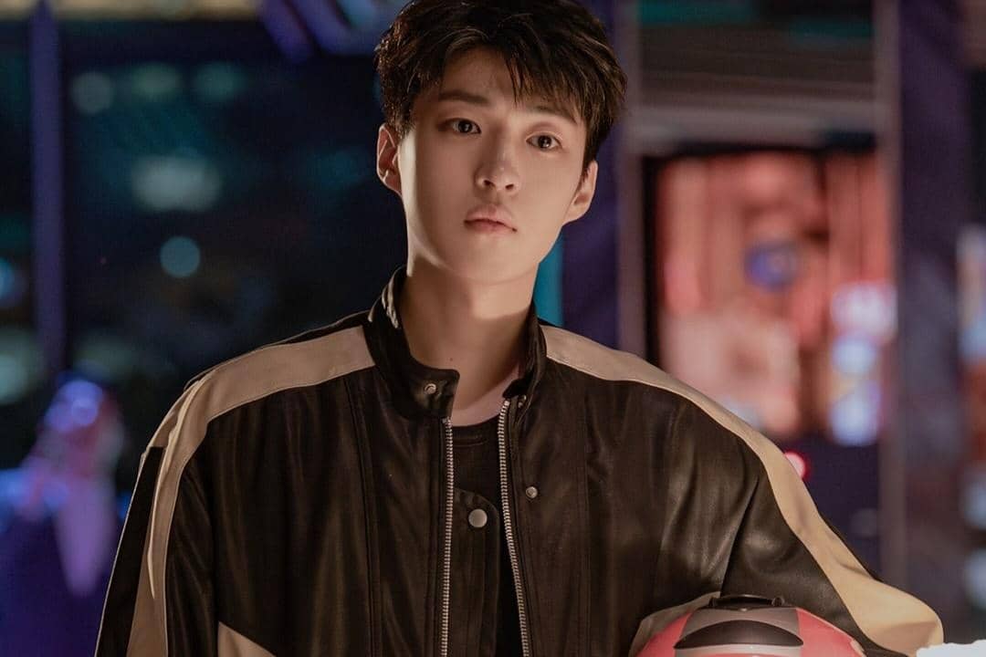 Seorang pria muda mengenakan jaket kulit hitam dan memegang helm merah dalam adegan drama Dream to You dengan latar malam berwarna neon.