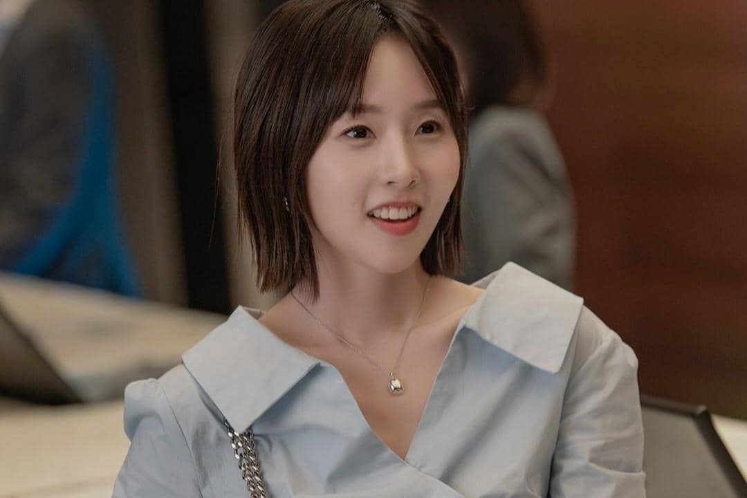 Seorang wanita berambut pendek mengenakan blus biru muda tersenyum hangat dalam adegan drama Dream to You.