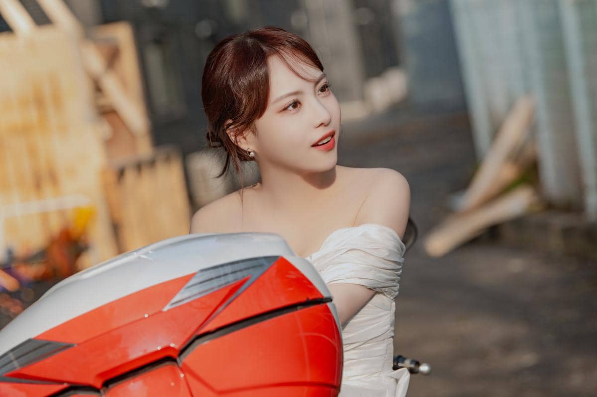 Seorang wanita mengenakan gaun putih duduk di atas motor berwarna merah dan putih di area luar ruangan dengan pencahayaan hangat.