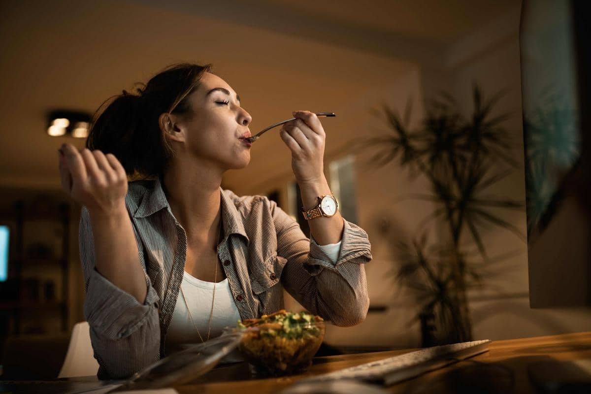 Seorang wanita duduk di meja makan sambil menikmati makanan dari mangkuk dengan sendok di tangan di suasana rumah yang hangat.