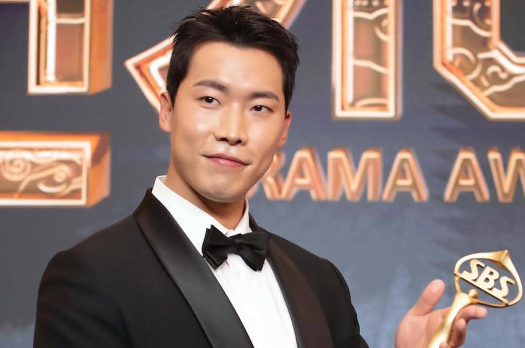 Seorang pria mengenakan setelan jas hitam dan dasi kupu-kupu memegang trofi bertuliskan SBS di acara penghargaan drama.