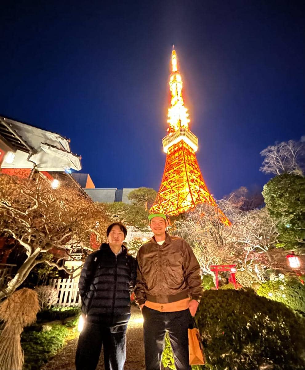 Dua pria berpose di malam hari dengan latar belakang Tokyo Tower yang menyala terang di tengah taman yang diterangi lampu.