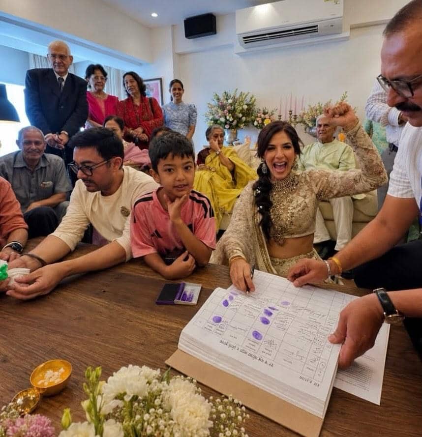Pasangan pengantin sedang menandatangani dokumen pernikahan di hadapan keluarga yang tersenyum dan bersuka cita di dalam ruangan.