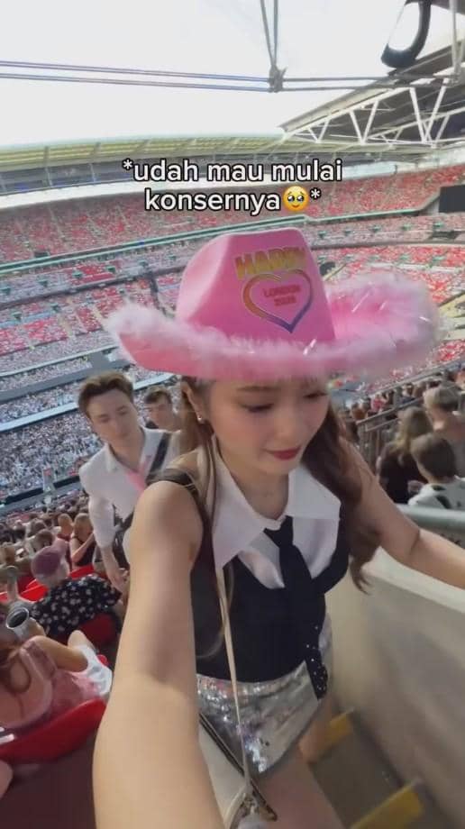Seorang perempuan mengenakan topi koboi pink dengan tulisan warna-warni dan pakaian pesta sedang menaiki tangga di stadion konser yang penuh penonton.
