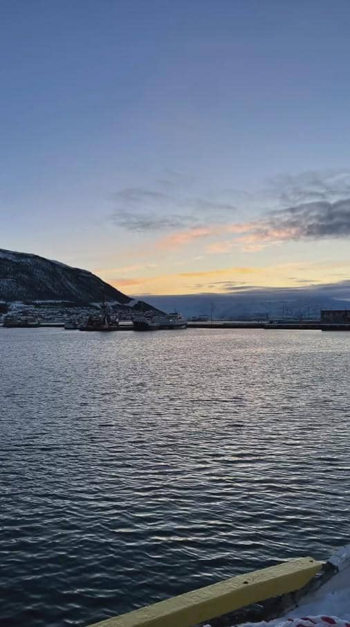 Pemandangan pelabuhan di Norwegia saat senja dengan air laut tenang, pegunungan bersalju, dan langit berwarna jingga kebiruan.