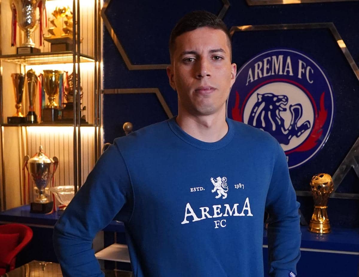 Seorang pria mengenakan sweater biru bertuliskan Arema FC berpose di depan logo klub dan deretan trofi di ruangan piala.