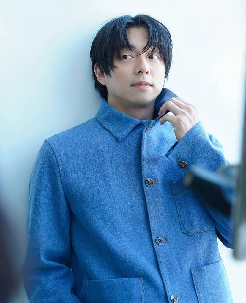 Seorang pria berambut hitam mengenakan jaket denim biru menatap ke samping sambil memegang kerah bajunya di latar putih.