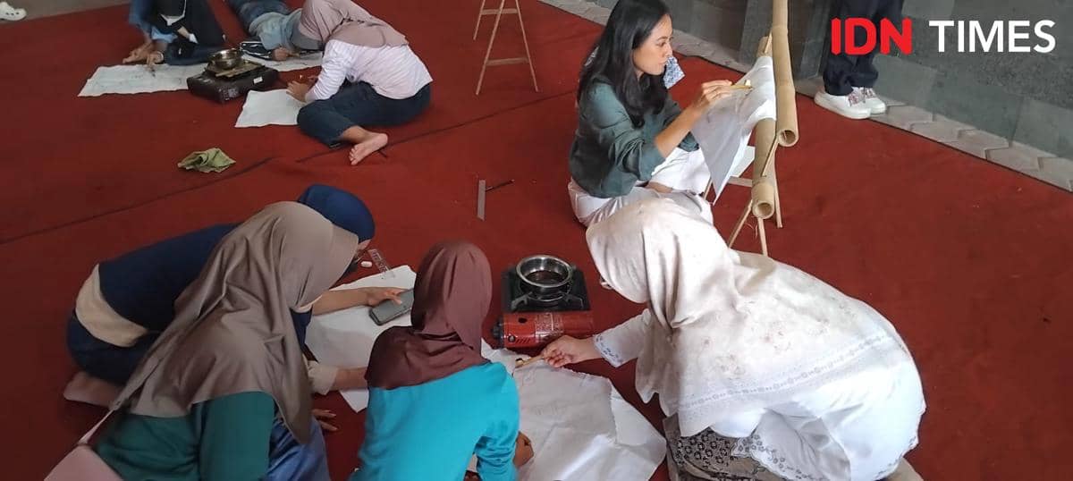 Beberapa perempuan duduk di lantai berkarpet merah sambil membuat batik dengan canting dan kain putih dalam kegiatan seni.