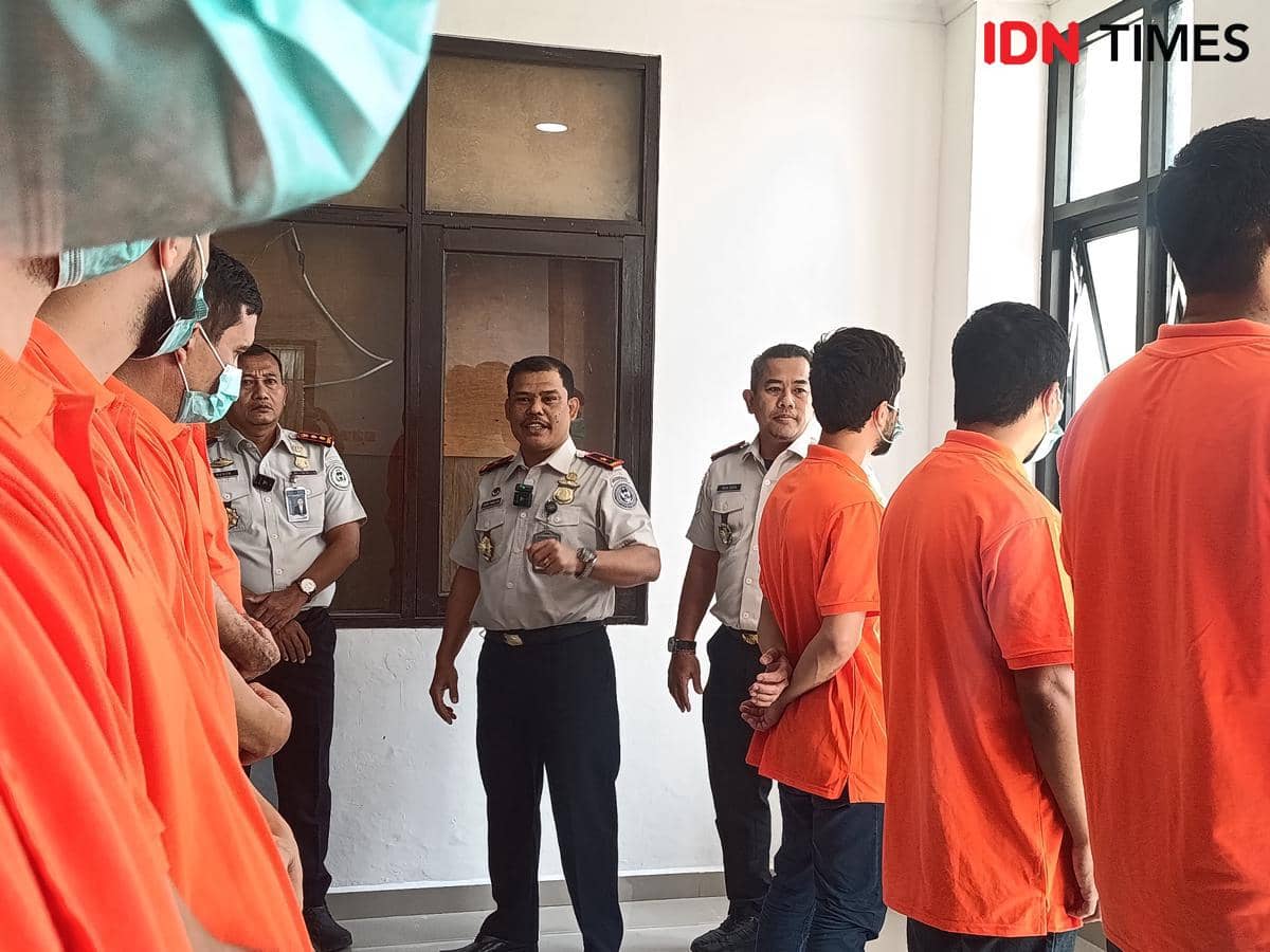 Petugas imigrasi memeriksa kondisi sejumlah warga asing berpakaian oranye di dalam ruangan dengan pencahayaan alami dari jendela.