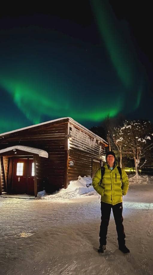 Seorang pria mengenakan jaket tebal berwarna hijau berdiri di depan bangunan kayu bersalju dengan latar cahaya aurora borealis di langit malam Norwegia.