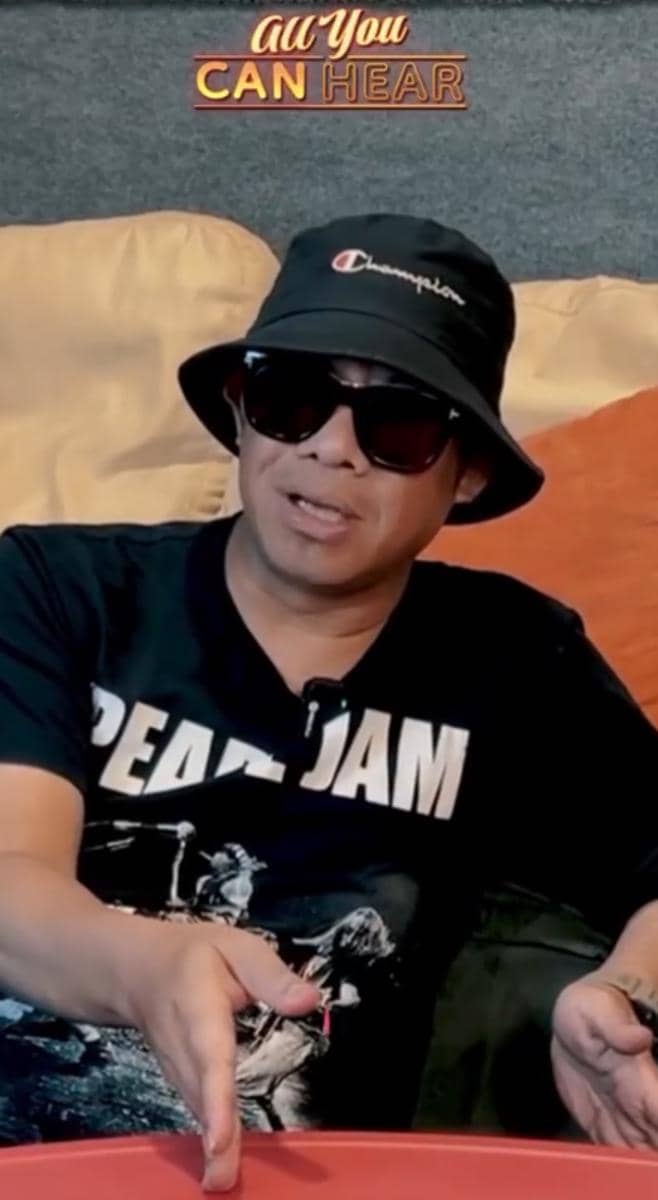 Seorang pria mengenakan topi bucket hitam, kacamata hitam, dan kaus bergambar band duduk di sofa dengan latar bertuliskan All You Can Hear.