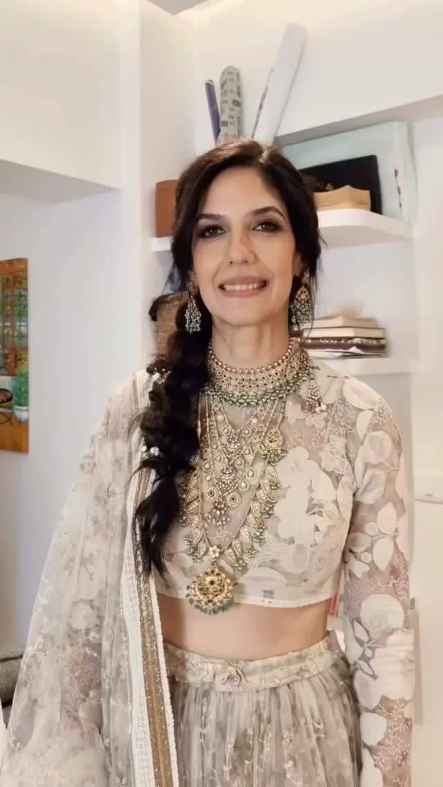 Seorang wanita mengenakan lehenga berwarna krem bermotif bunga dengan perhiasan emas tradisional dan rambut dikepang di dalam ruangan.