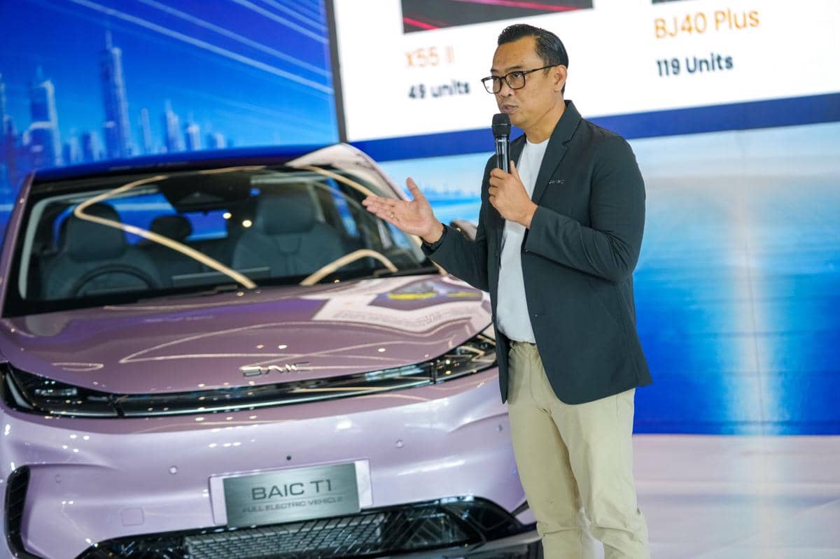 Seorang pria berbicara dengan mikrofon di samping mobil listrik BAIC T1 berwarna ungu muda dalam sebuah acara presentasi otomotif.