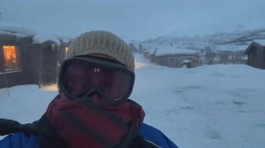 Seseorang mengenakan kacamata salju, topi rajut, dan syal tebal berdiri di area bersalju dengan latar pegunungan dan rumah kayu di Norwegia.